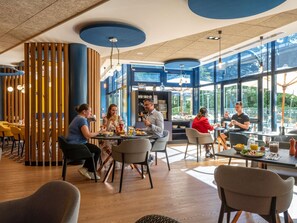 A modern dining area with round tables, wooden partitions, and large windows offering views of greenery.