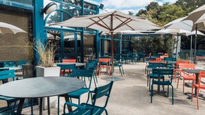 Un café en plein air avec des tables et des chaises, des parasols, et un bâtiment bleu en arrière-plan.