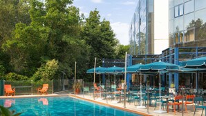 A swimming pool with lounge chairs and umbrellas, surrounded by greenery and a modern building. - Novotel La Rochelle Centre (La Rochelle)