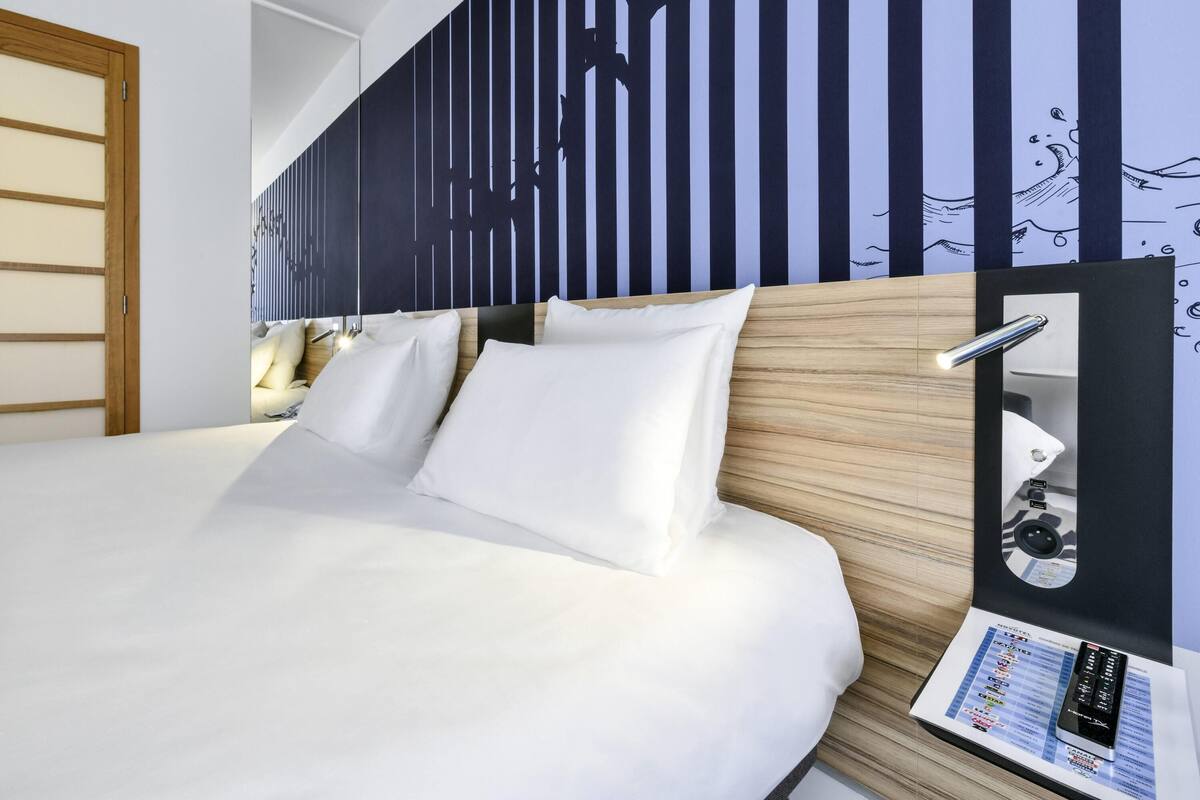 Una habitación de hotel con una cama grande, un cabecero de madera, un espejo fijado a la pared y una mesita de noche con una lámpara.