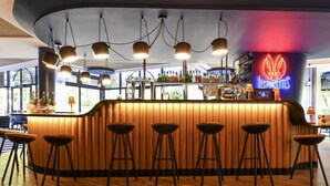 A bar area with a neon sign, wooden bar counter, stools, and a view through large windows. - Novotel La Rochelle Centre (La Rochelle)
