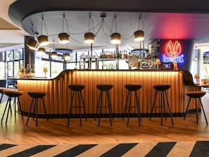 A bar area with a neon sign, wooden bar counter, stools, and a view through large windows.