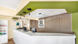 A hotel reception area with a white reception desk, a 'Quality by Choice Hotels' sign, a 'Welcome' sign, and a green accent wall. - Quality Inn Klamath Falls - Crater Lake Gateway (Klamath Falls)