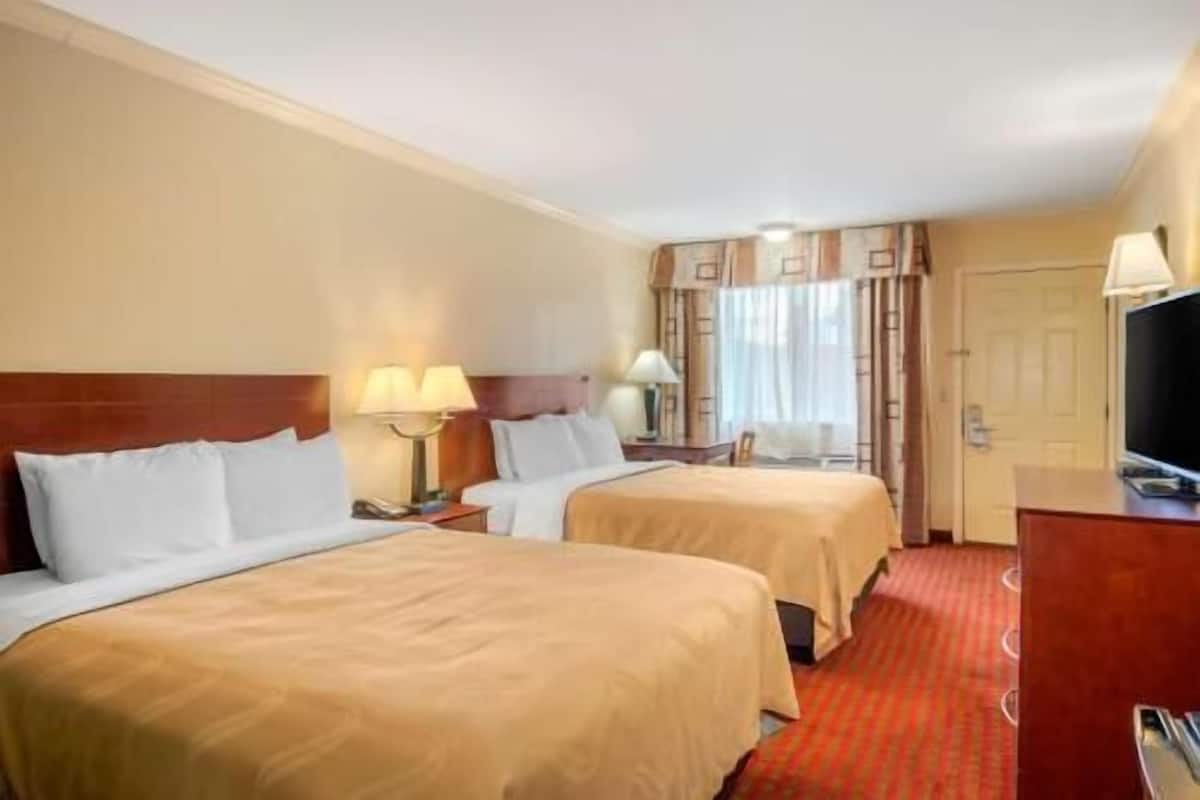 A hotel room with two beds, a television, a desk, and a window with curtains.