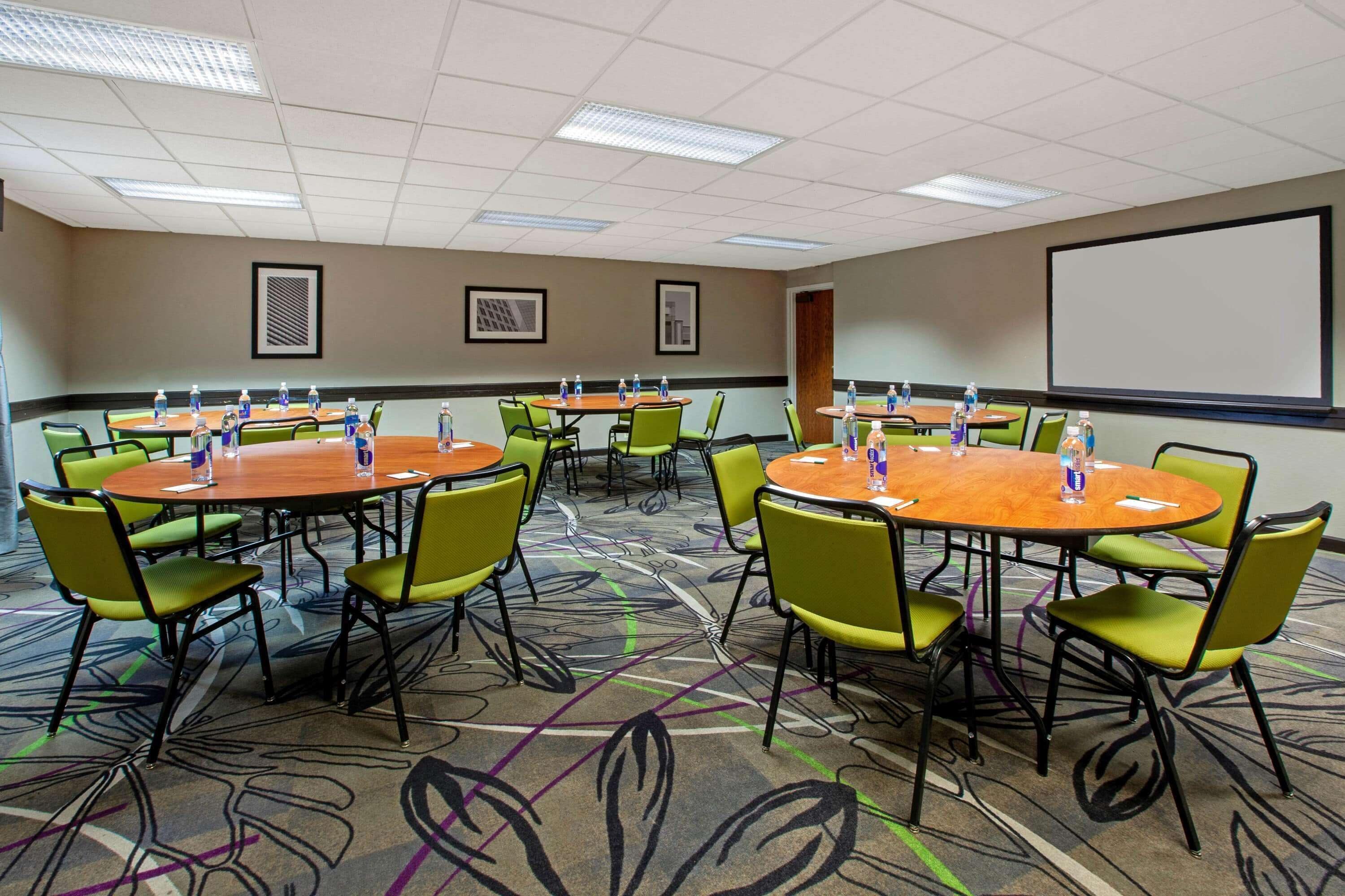 A conference room with multiple tables and chairs, a projector screen, and framed artwork on the walls.