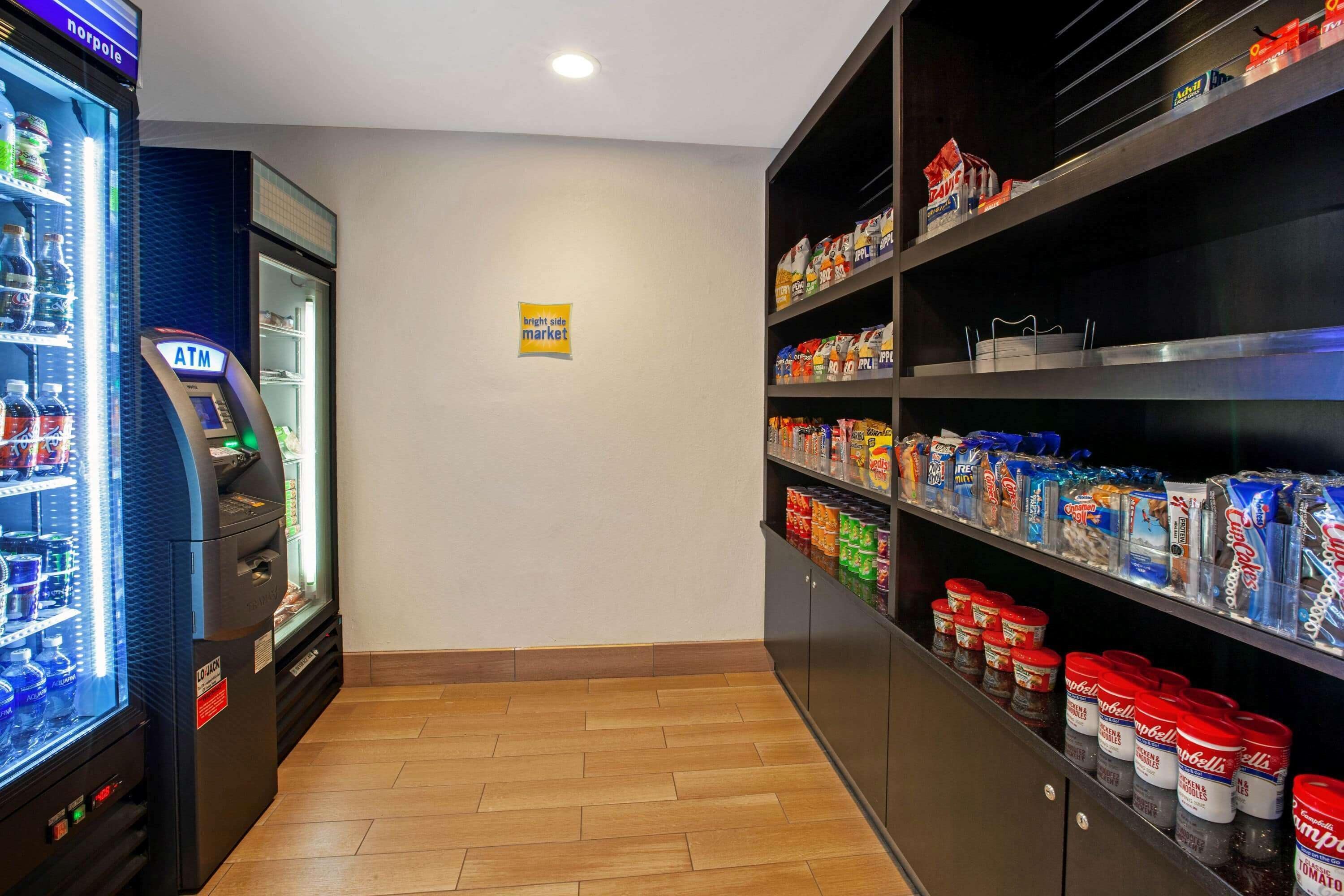 A self-service kiosk with an ATM, a refrigerated beverage dispenser, and shelves stocked with snacks and condiments.