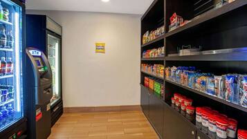 A self-service kiosk with an ATM, a refrigerated beverage dispenser, and shelves stocked with snacks and condiments.