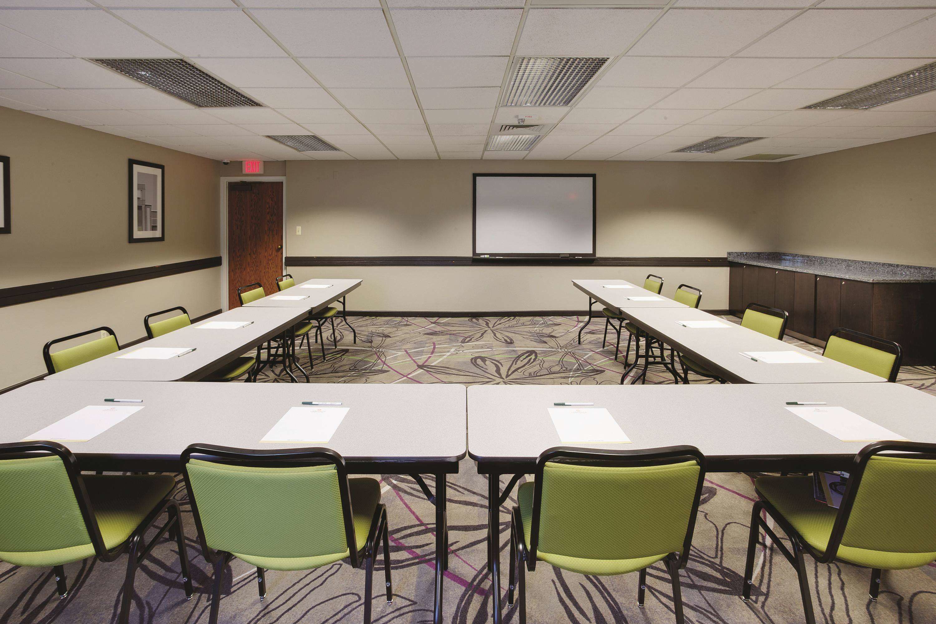 A conference room with a long table, green chairs, a projector screen, and a carpeted floor.