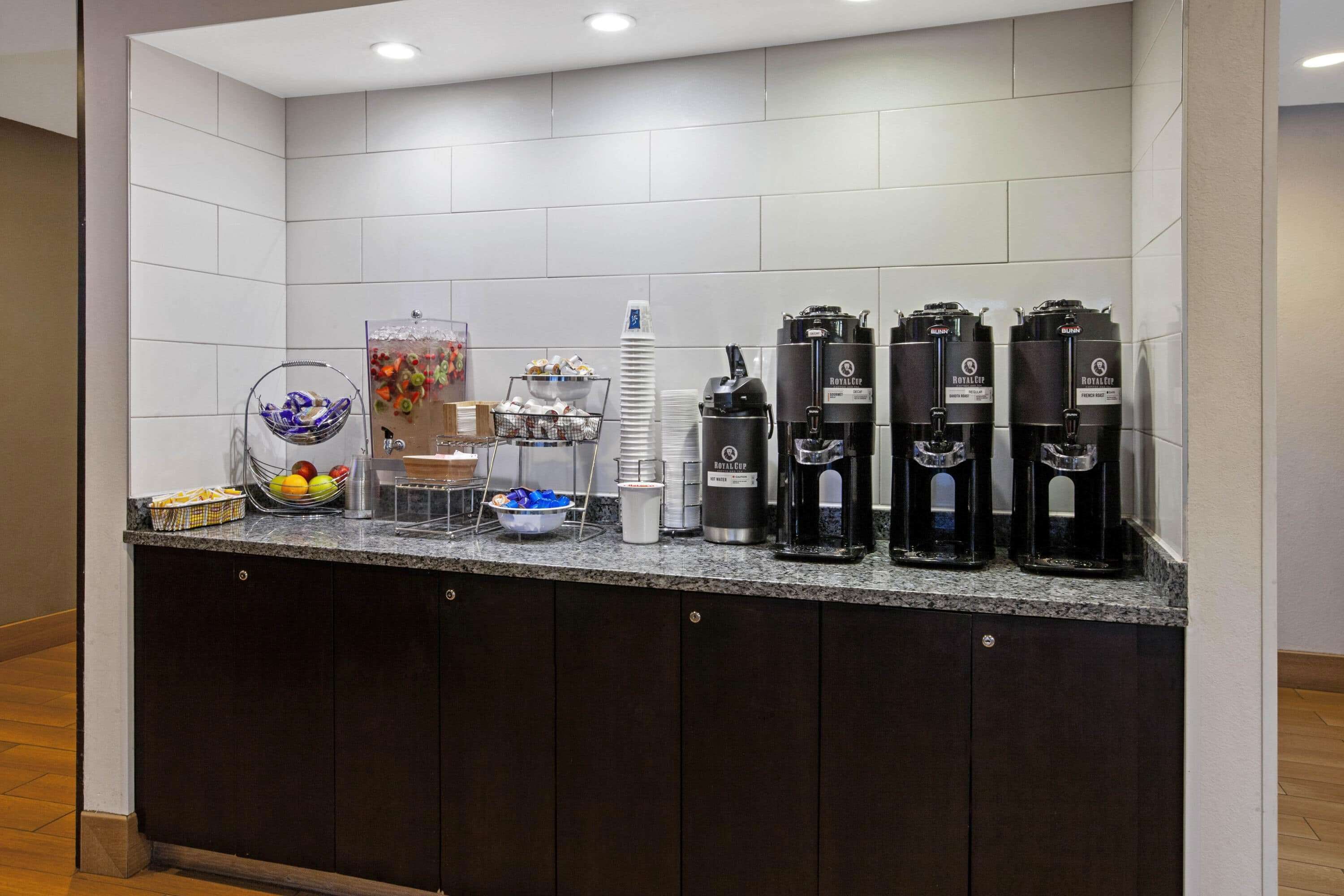 A self-service beverage station with multiple coffee machines, a fruit bowl, and a basket of snacks.