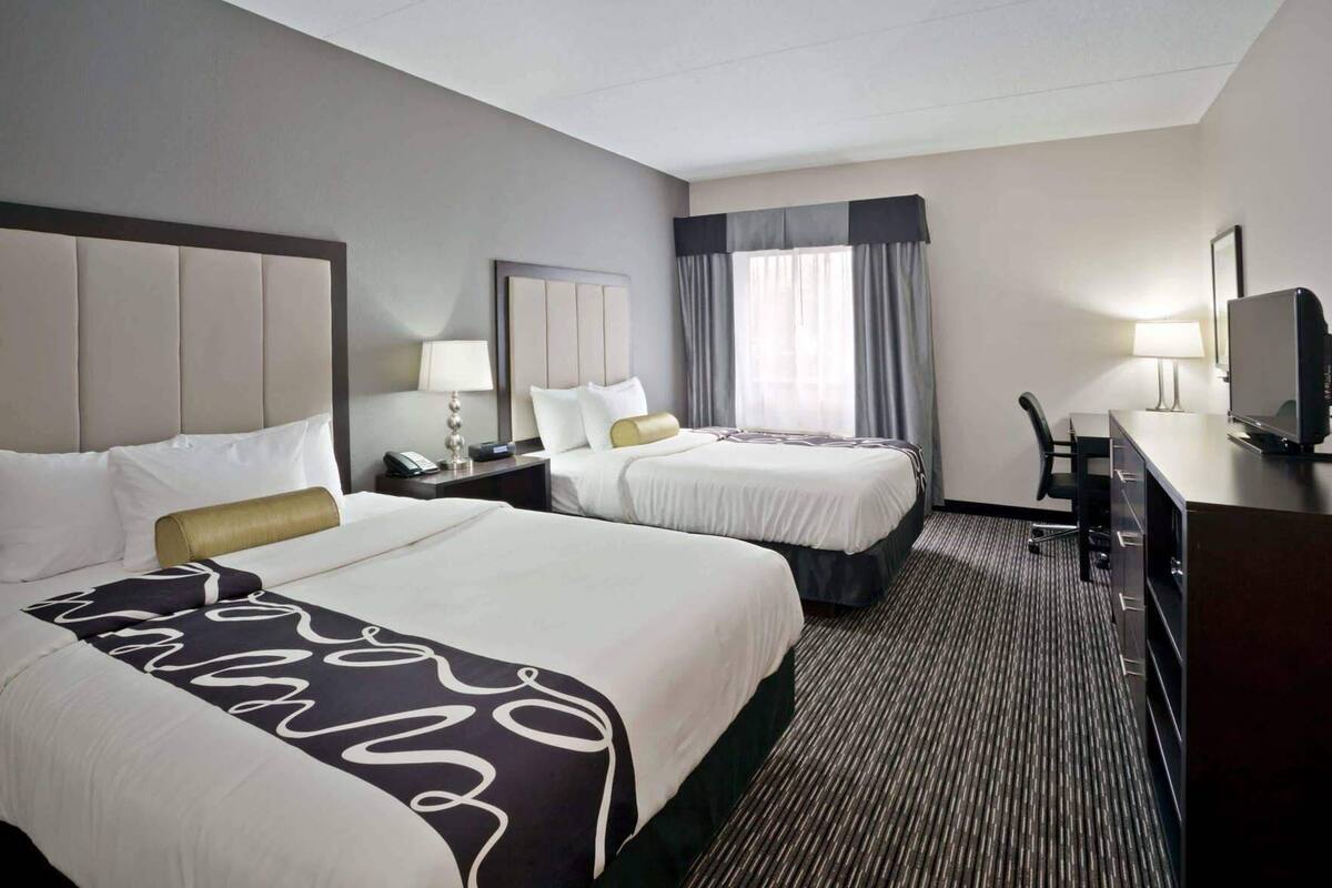 Ein Hotelzimmer mit zwei Betten, einem Schreibtisch mit Fernseher, einem Sessel, einer Lampe und einem Fenster mit Vorhängen.