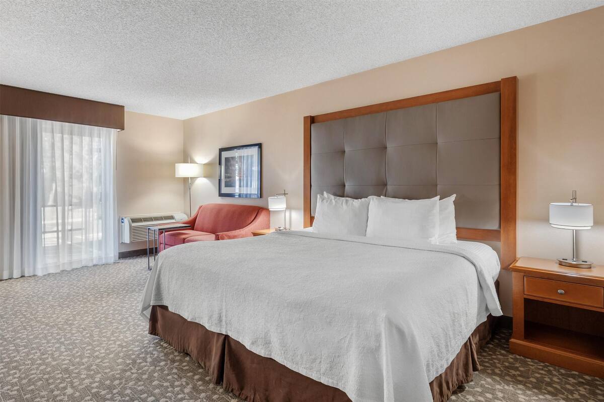 A hotel room with a large bed, a red chair, a nightstand, and a window with curtains.
