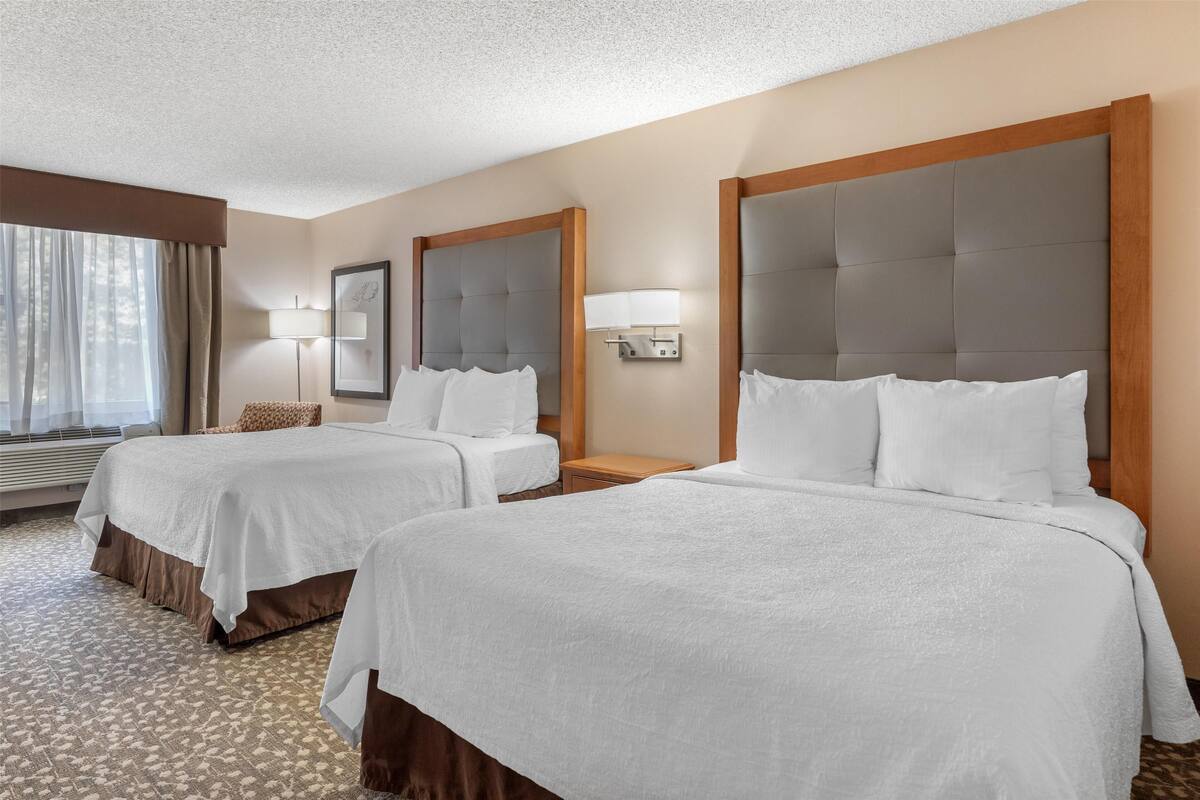 A hotel room with two beds, a large window with curtains, a bedside table with a lamp, and a wall-mounted light fixture.