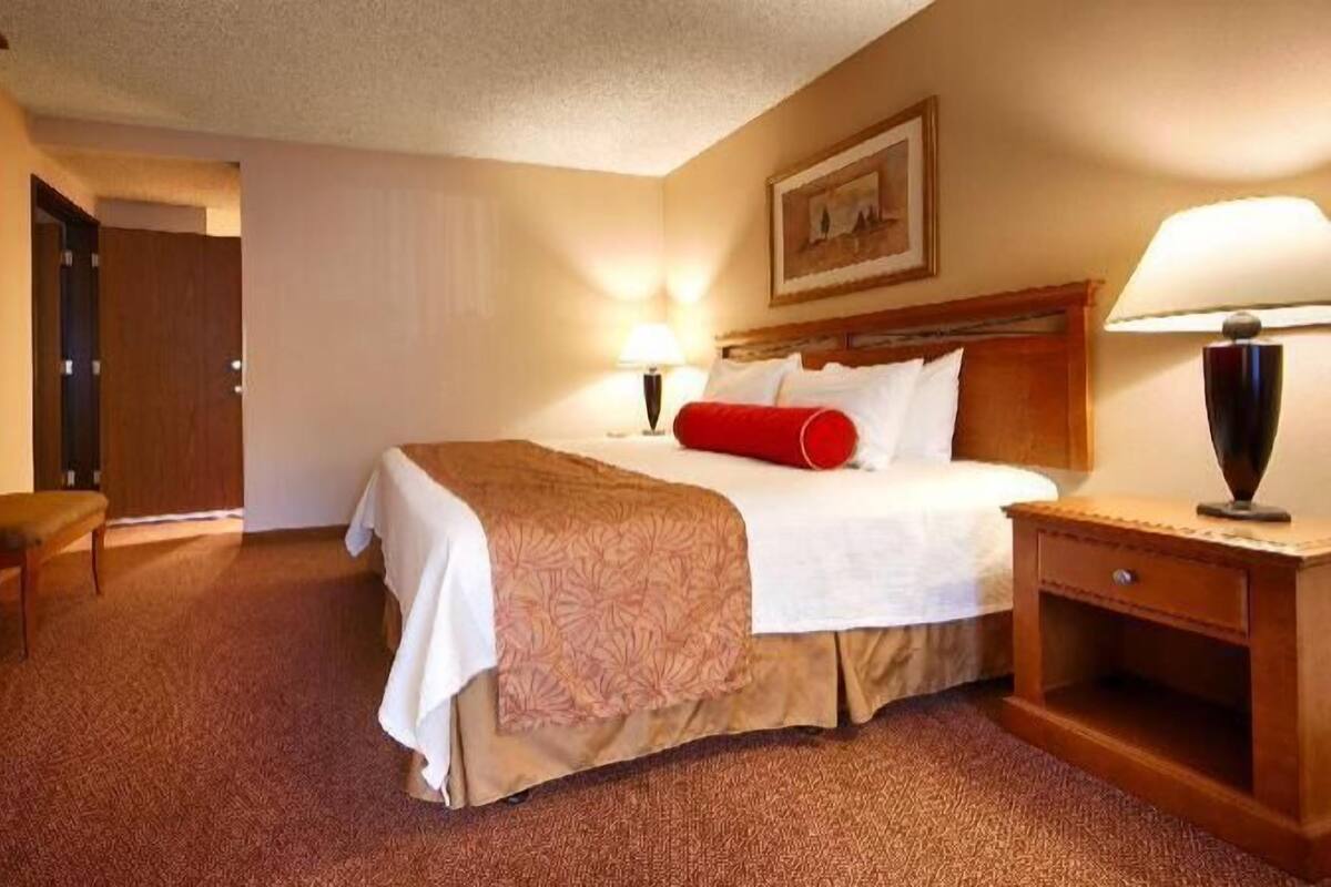 A hotel room with a bed, bedside table, lamp, and a door leading to another room.