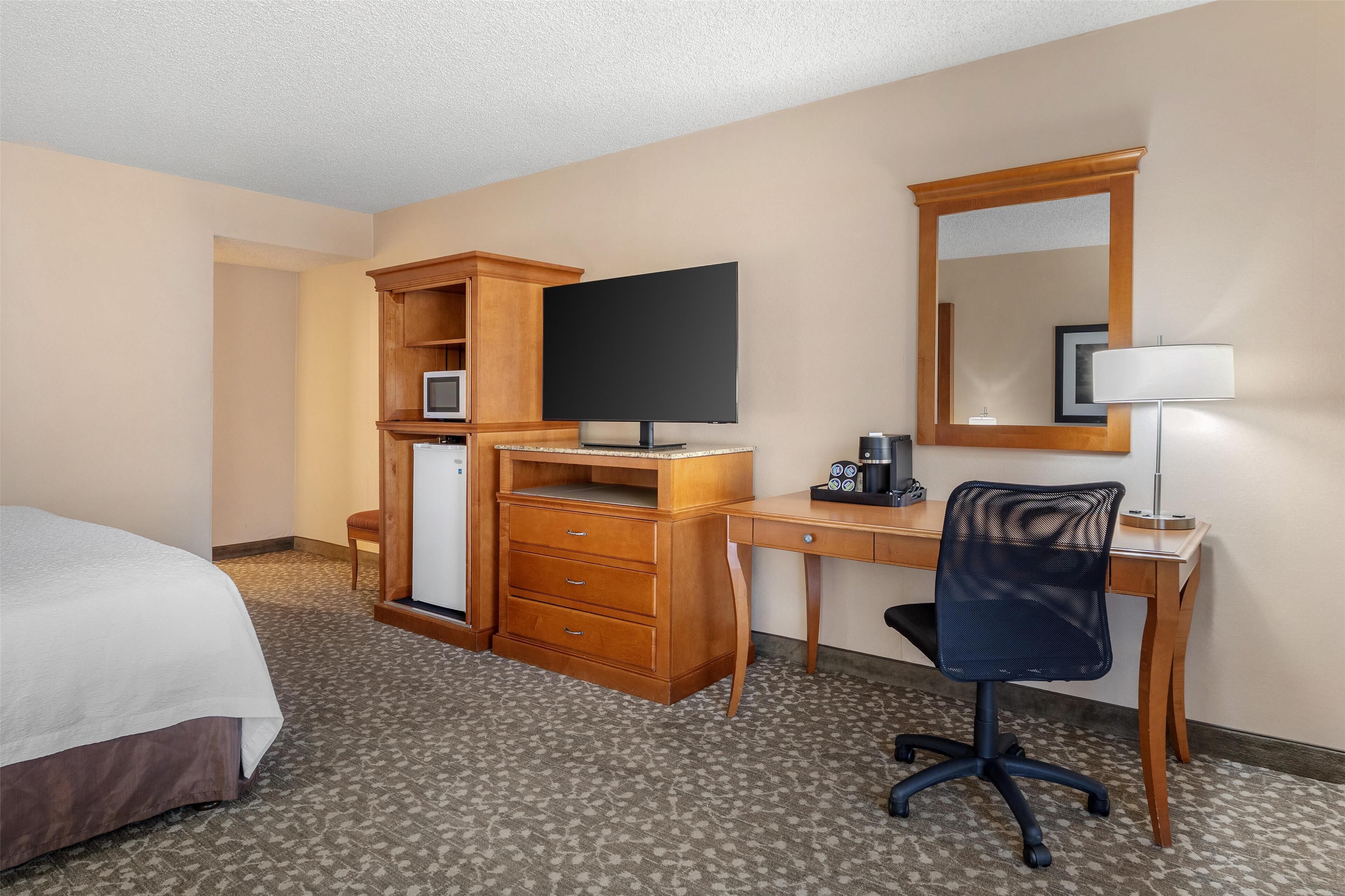 A hotel room with a bed, a desk with a computer, a chair, a TV, a mirror, and a lamp.