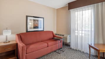 Habitación de hotel con un sofá rojo, una mesita, una lámpara, un cuadro en la pared y una ventana con cortinas translúcidas.