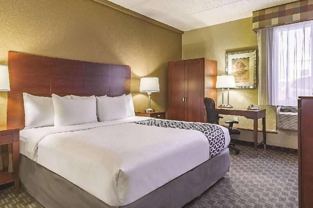 Habitación de hotel con una cama grande, un cabecero de madera, un escritorio con silla, un armario y una ventana con cortinas.