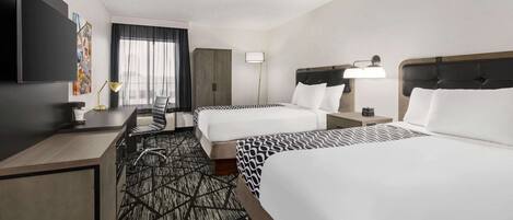 A hotel room with two beds, a desk, a TV, and a large window with curtains.