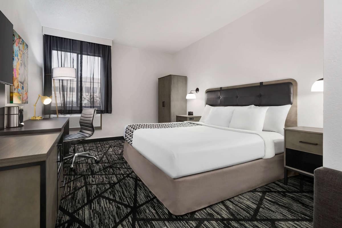 Una habitación de hotel con una cama grande, un escritorio con lámpara, una silla, un armario y una ventana con cortinas.