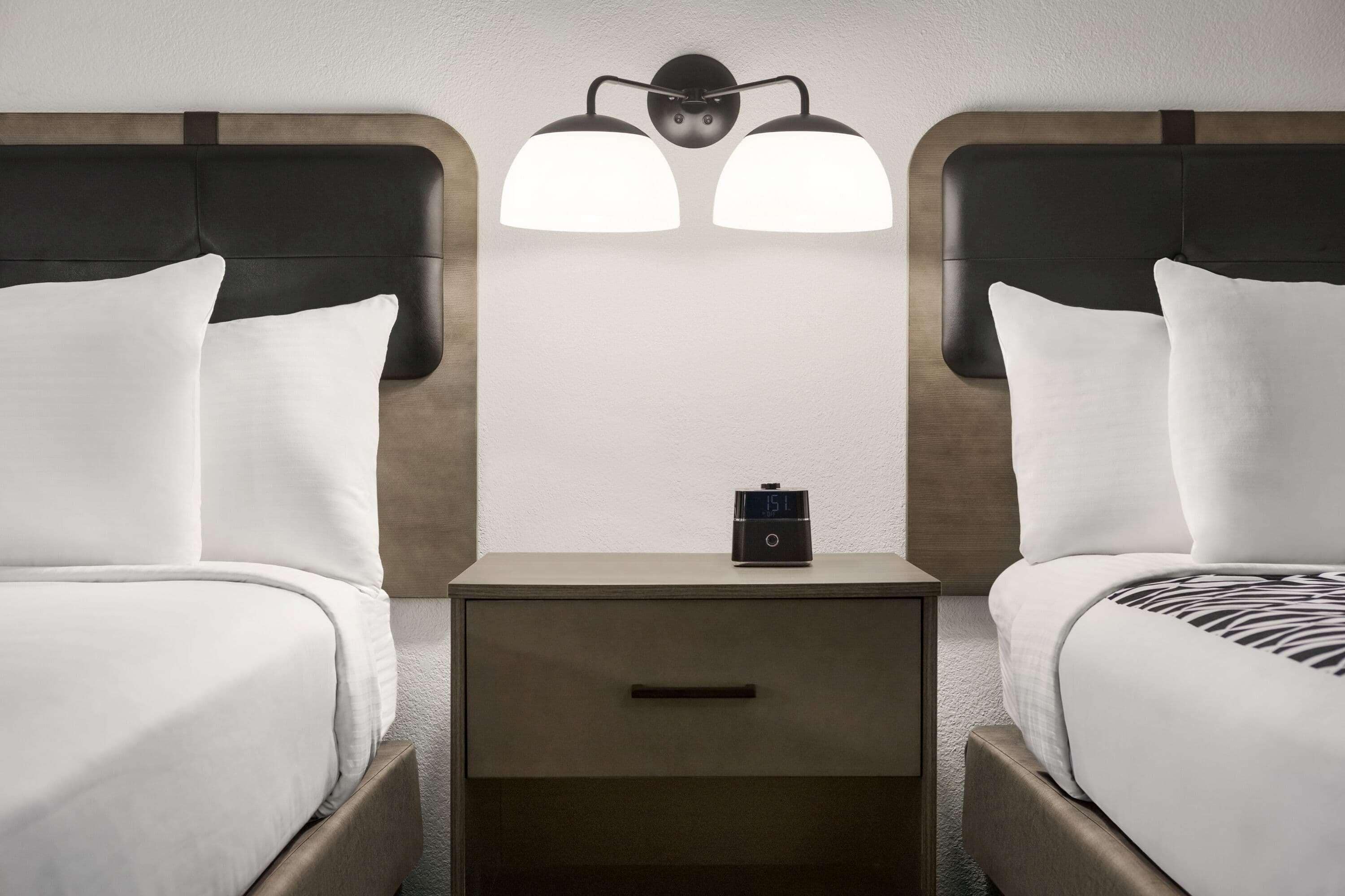 Une chambre d’hôtel avec deux lits, une table de chevet avec une horloge et un luminaire fixé au mur.