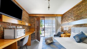 Quarto de hotel com parede de tijolos, televisão de tela plana, micro-ondas, cama com colcha estampada em azul e branco, e mesa de trabalho com cadeira.