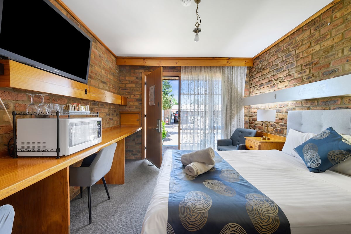 Habitación de hotel con pared de ladrillo, televisor de pantalla plana, microondas, cama con colcha de diseño azul y blanco, y escritorio con silla.