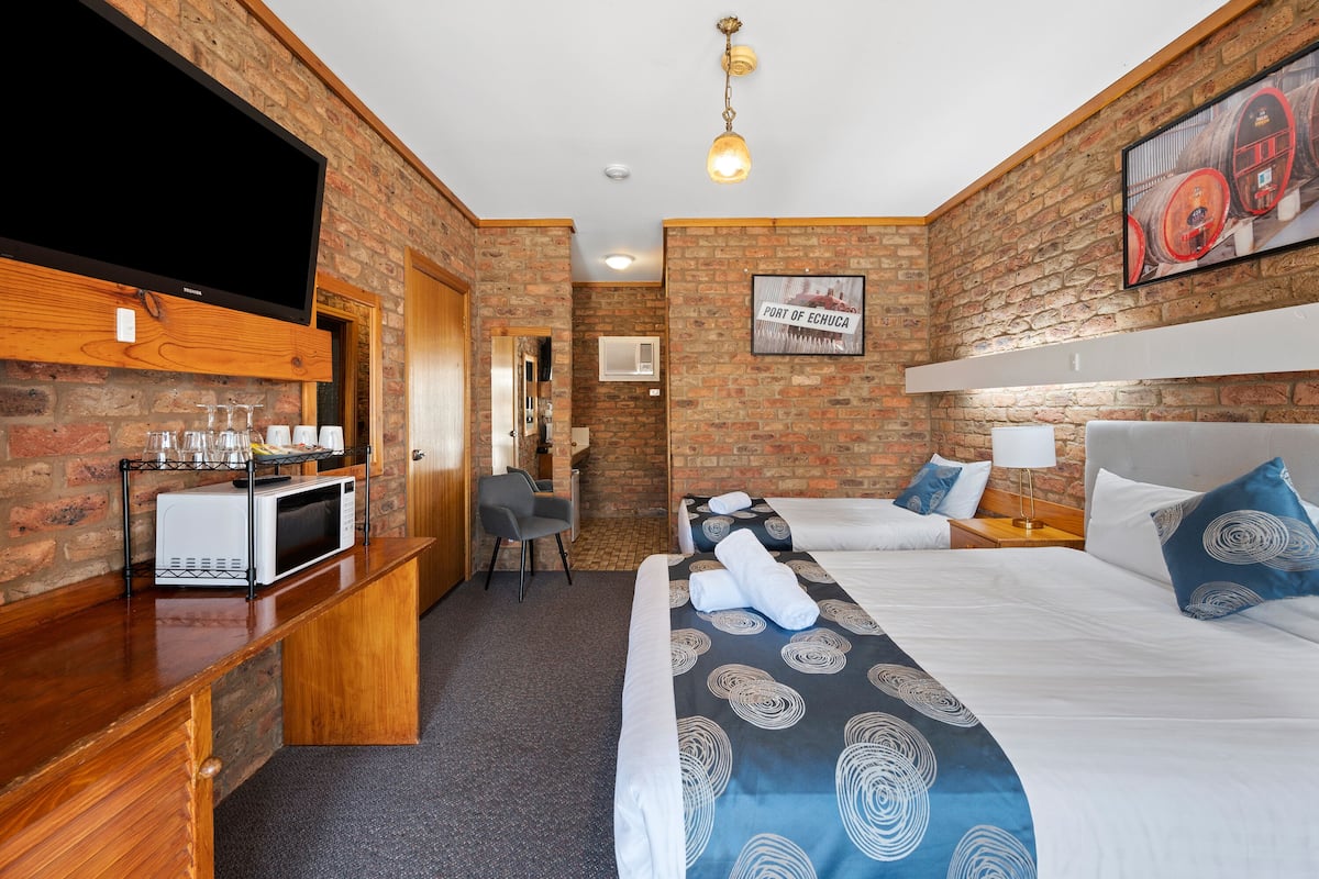 Habitación de hotel con pared de ladrillo, un televisor de pantalla plana, microondas, cama con ropa de cama azul y blanca, y un pequeño escritorio con una silla.