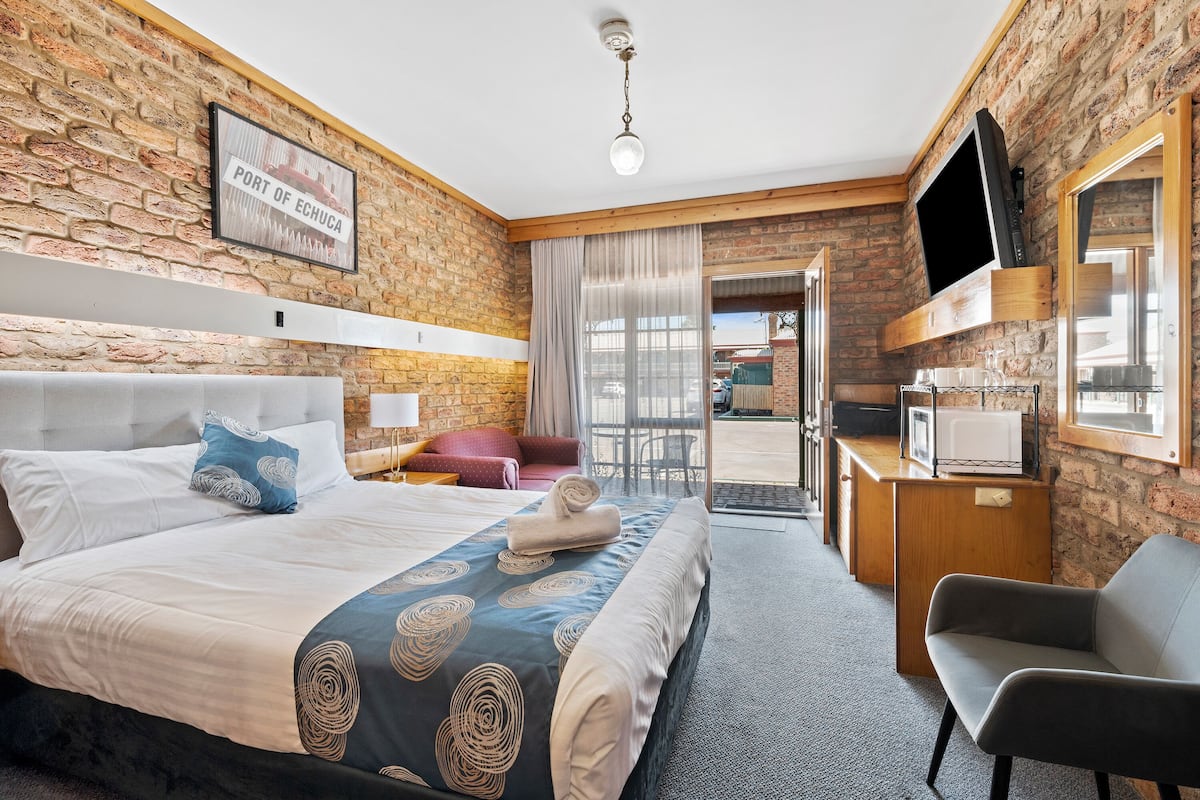 Habitación de hotel con pared de ladrillo, cama con colcha de diseño azul y blanco, una silla gris y un televisor de pantalla plana montado en la pared.