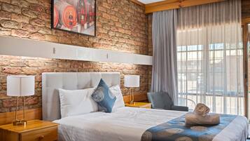 A hotel room with a brick wall, a large bed with a blue and white patterned bedspread, two bedside lamps, a wooden nightstand, a grey chair, and a large window with curtains.
