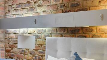 Una cama bien hecha con ropa blanca y un almohadón azul con diseños circulares, frente a una pared de ladrillo y un cabecero de madera.