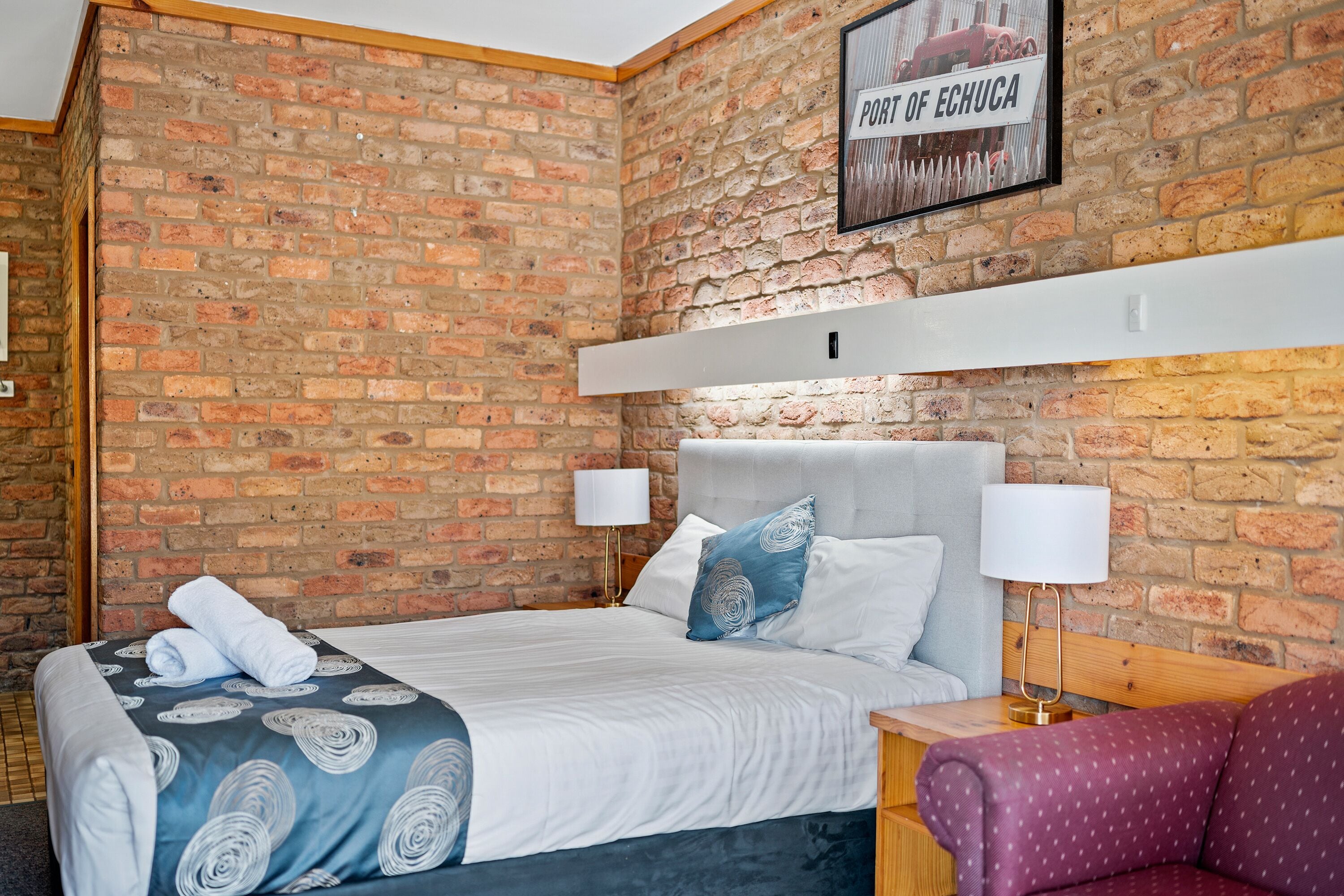 A hotel room with a brick wall, a bed with a blue and white patterned bedspread, two bedside lamps, and a framed picture on the wall.