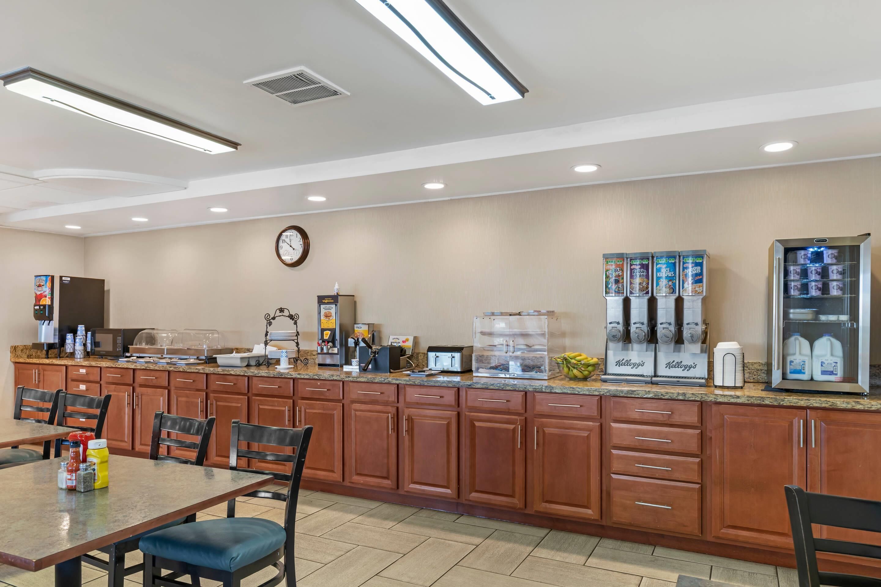 A hotel kitchen with a buffet setup, including a coffee machine, a refrigerator with milk, and a clock on the wall.