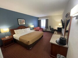 Habitación de hotel con una cama grande, un escritorio con computadora, una silla, una mesita con cafetera y un espejo.