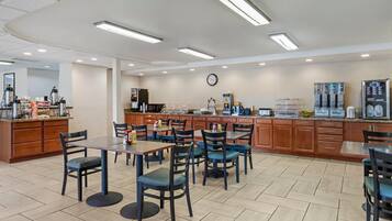 Un grand espace dédié à la salle à manger, avec plusieurs tables et chaises, un coin cuisine équipé d’appareils électroménagers et une horloge au mur.