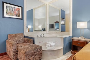 A hotel room with a large mirror, a sofa, a bed, a bedside table, a lamp, and a bathroom with a sink and towel rack.