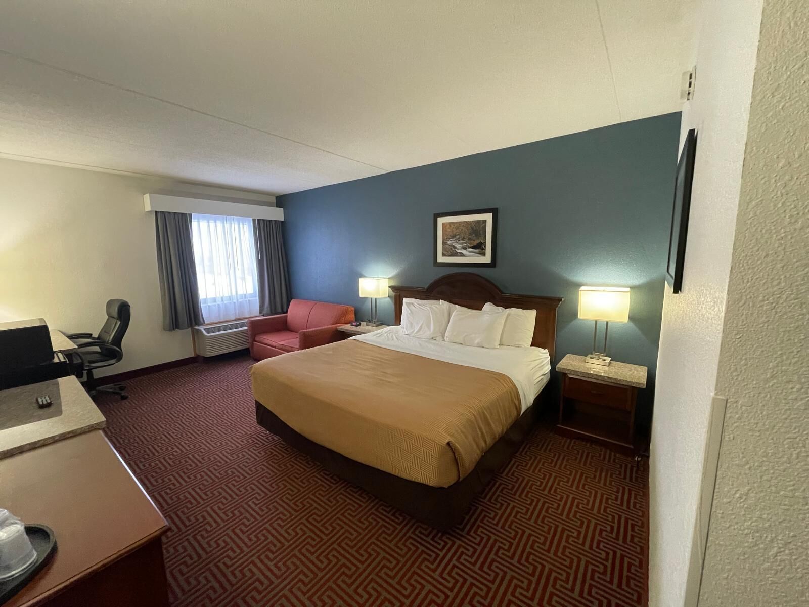 A hotel room with a large bed, a desk with a computer, a chair, and a window with curtains.