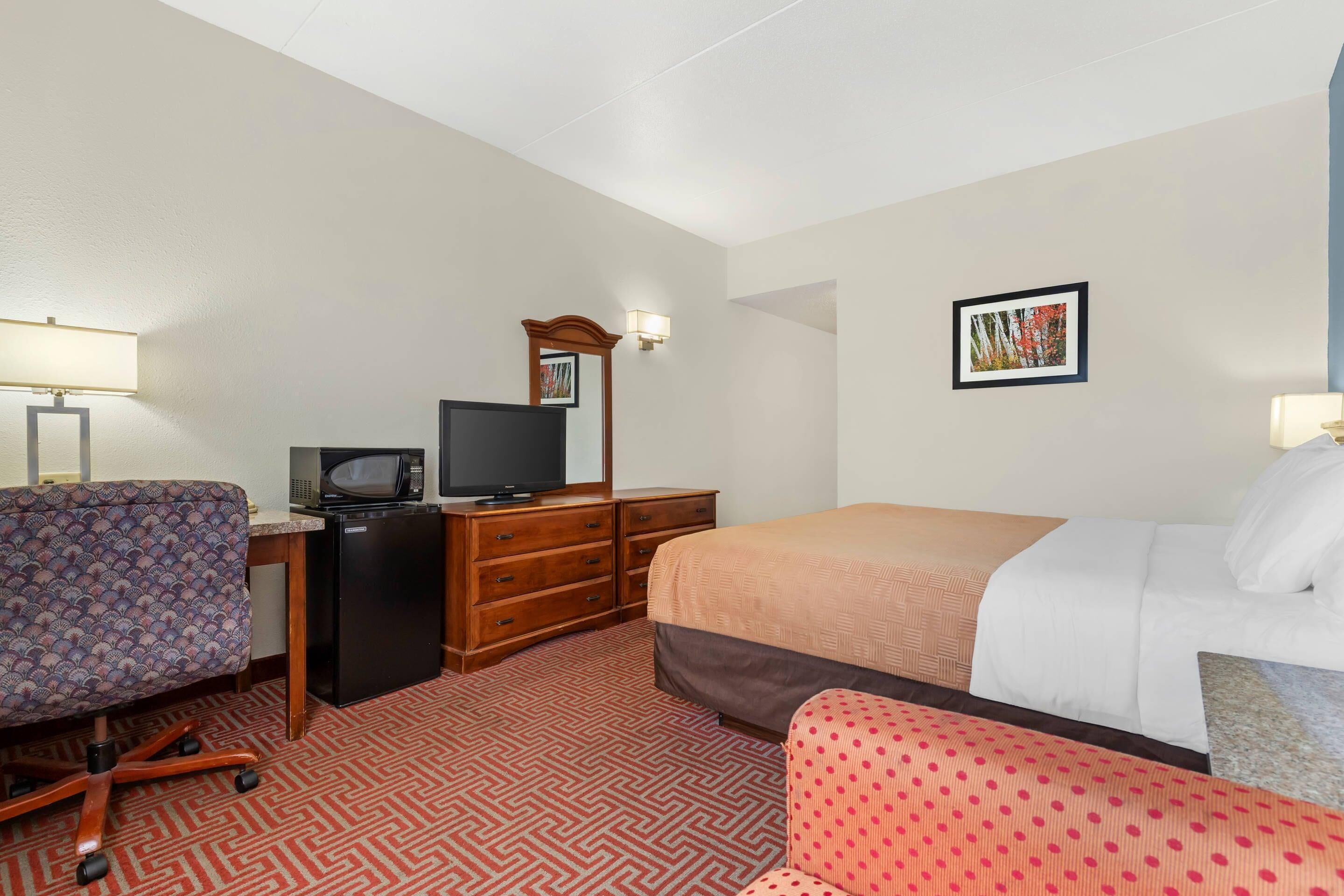 A hotel room with a bed, a desk with a television, a chair, a dresser, and a wall-mounted light.
