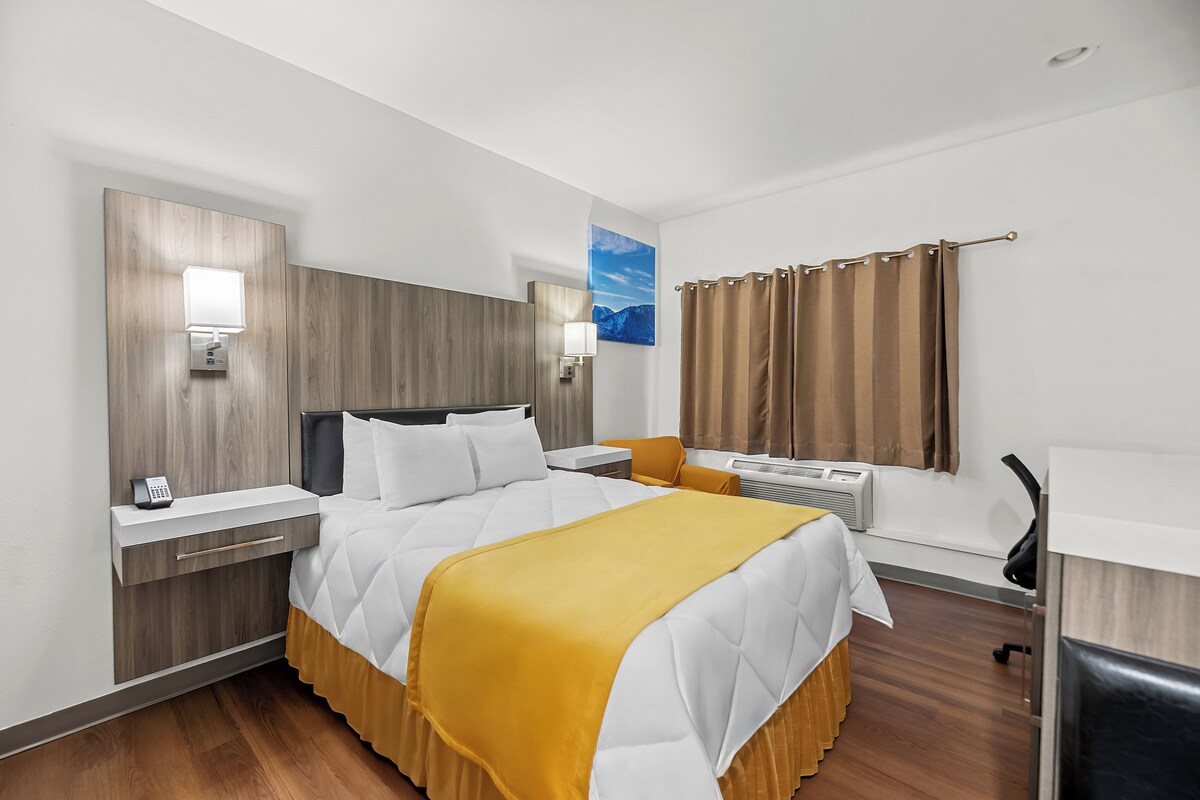 A hotel room with a bed, bedside tables, a desk, a chair, a window with curtains, and a painting on the wall.