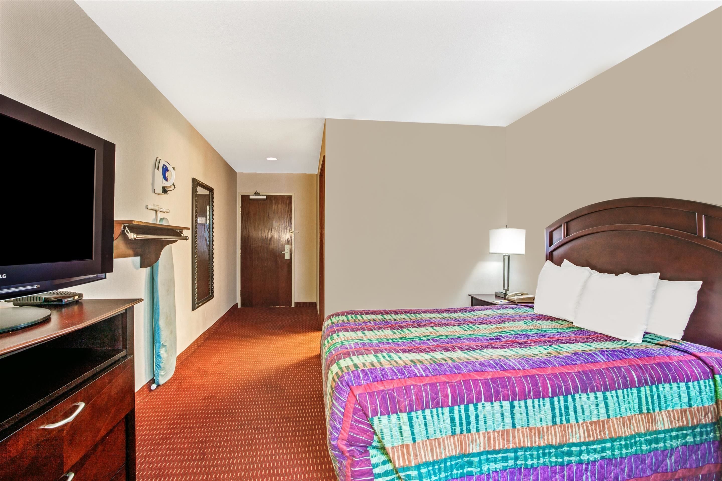 A hotel room with a large bed, a television on a dresser, a door leading to another room, and a nightstand with a lamp.