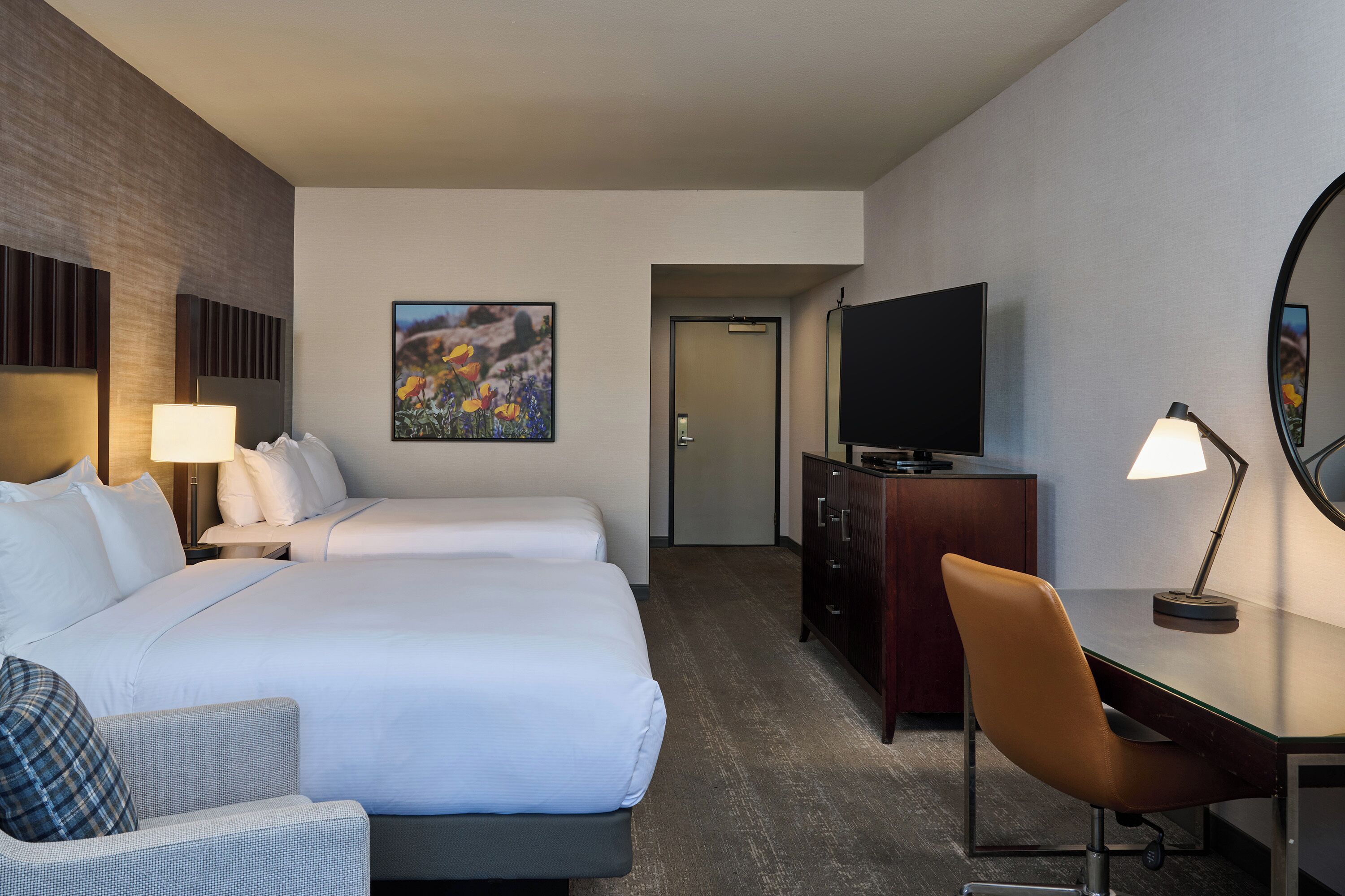 A hotel room with two beds, a desk, a chair, a TV, a dresser, and a painting on the wall.
