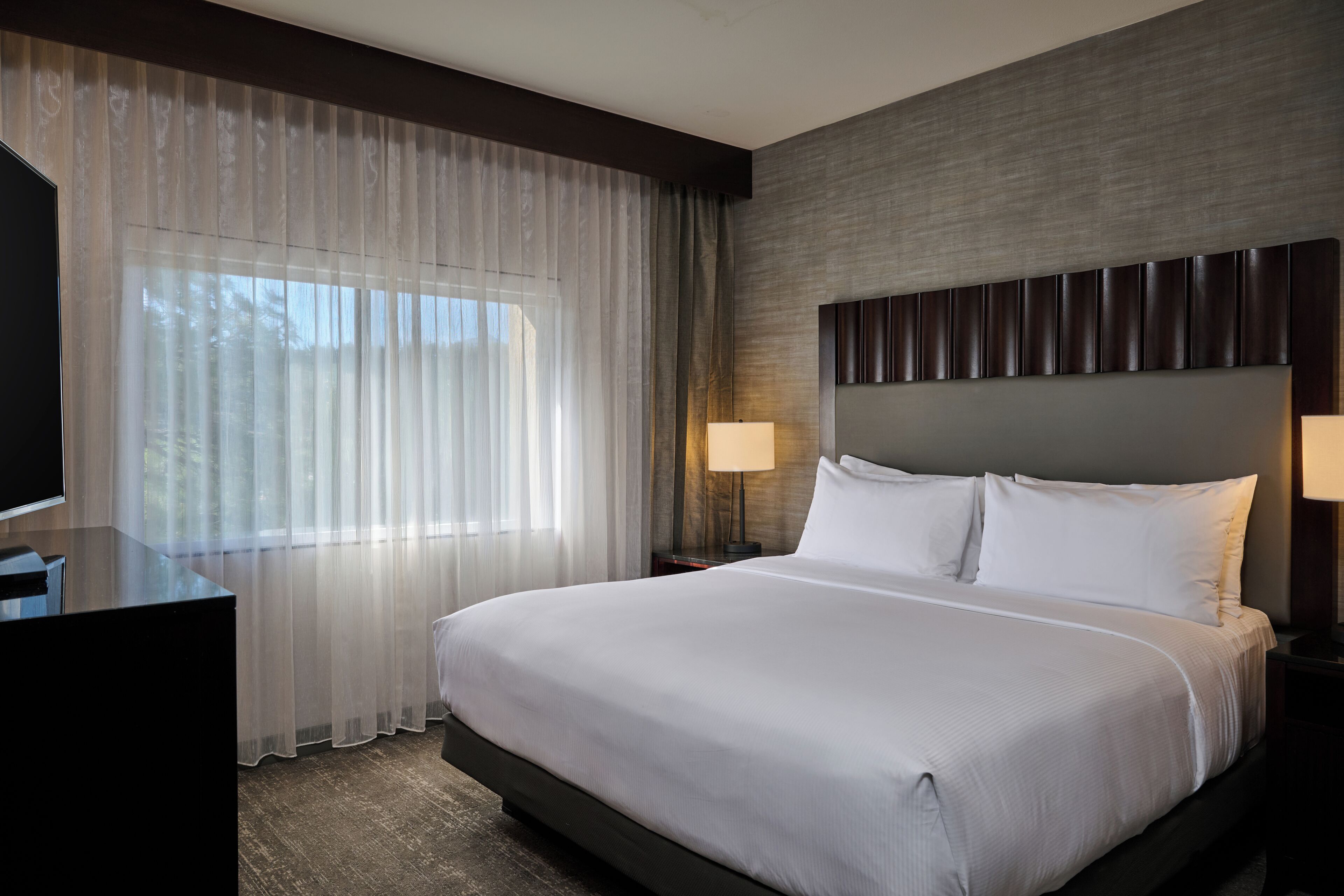 A hotel room with a large bed, a TV, a desk, and a window with sheer curtains.