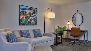 A hotel room with a grey sofa, a small table with a lamp, a desk with a chair, and a large framed picture on the wall.