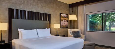 A hotel room with a large bed, a chair, a nightstand, a lamp, and a window with a view of trees.