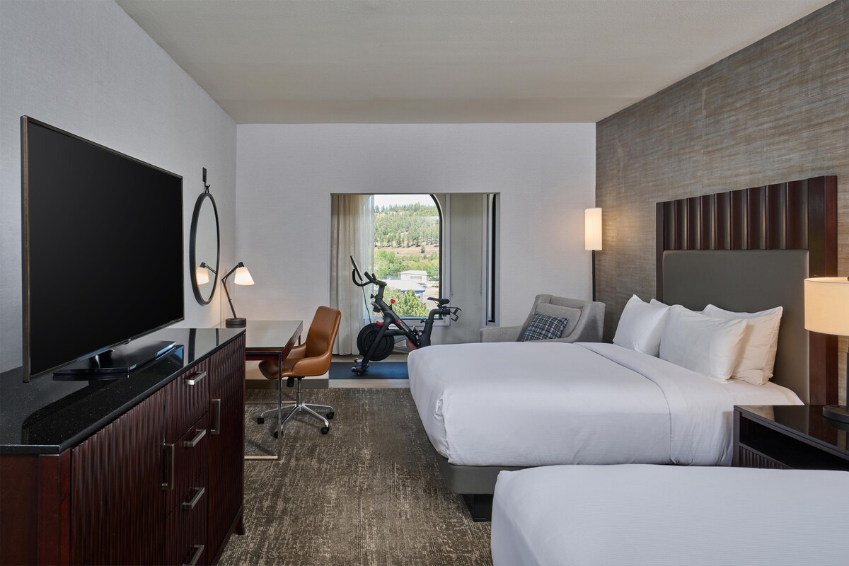A hotel room with a TV, exercise bike, two beds, a desk, and a view of greenery.