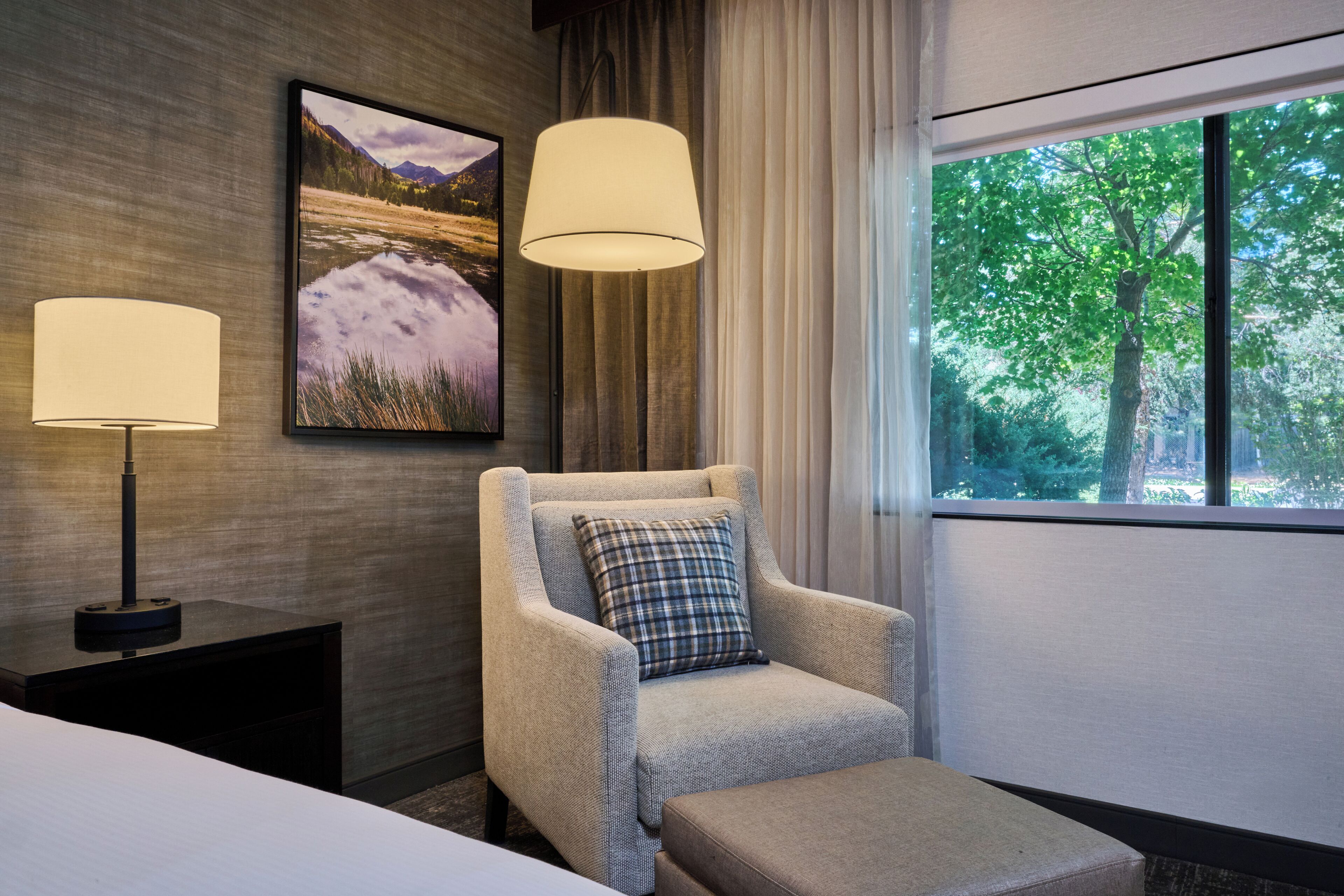 A hotel room with a bed, a chair with a pillow, a standing lamp, a framed picture, and a window with a view of trees.