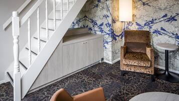 A room with a staircase, patterned carpet, a patterned armchair, a round table, and a lamp.