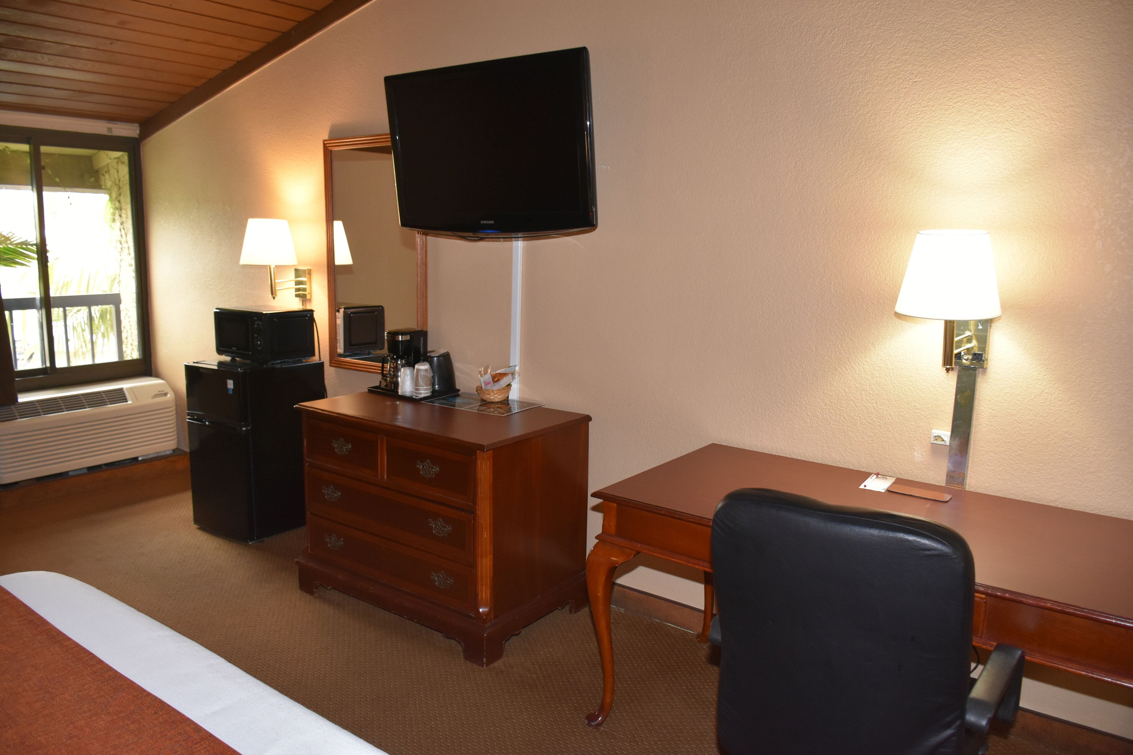 A hotel room with a bed, a wooden desk, a television, a dresser, and two wall-mounted lamps.