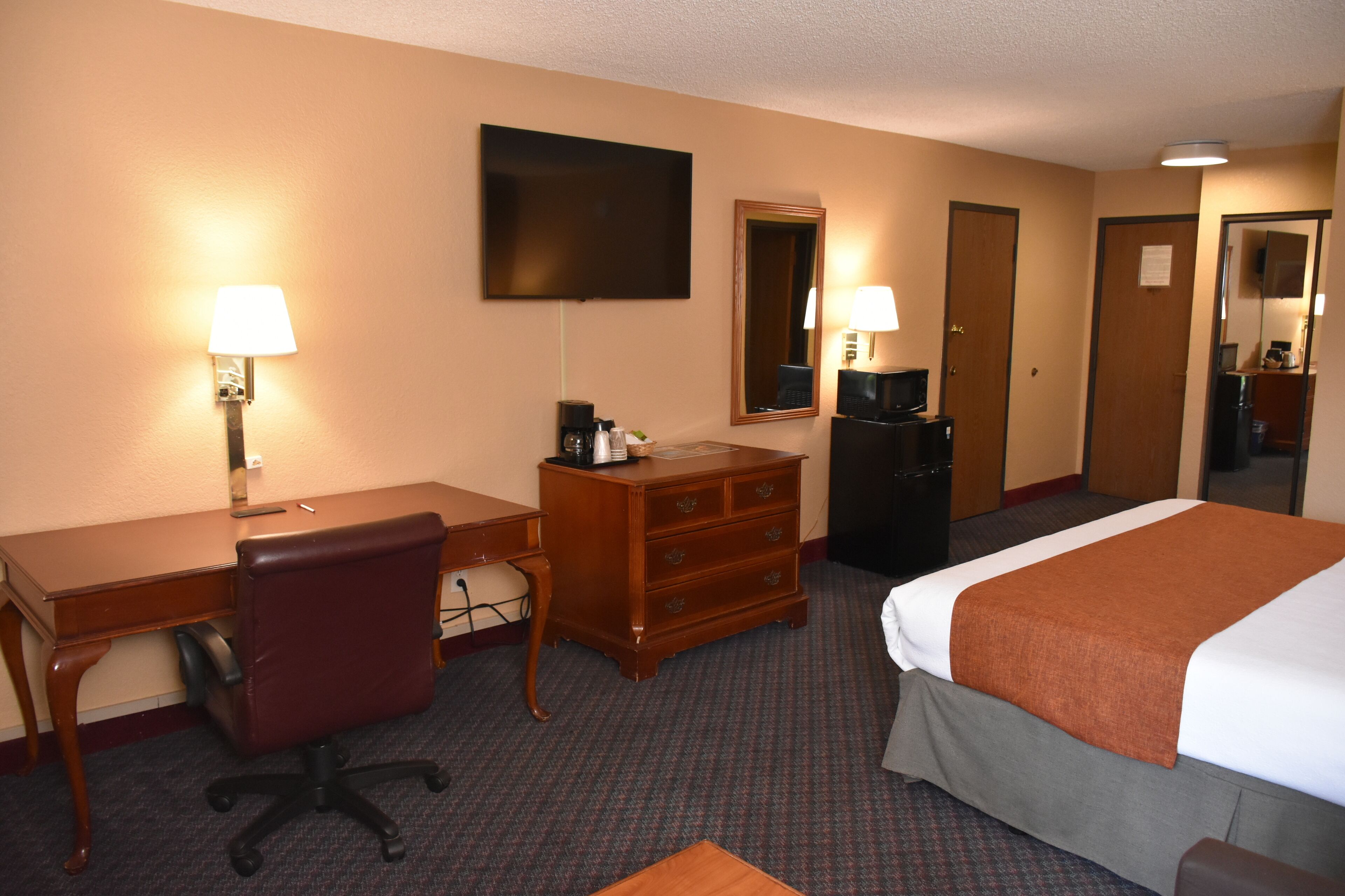A hotel room with a desk, chair, dresser, bed, and a TV mounted on the wall.