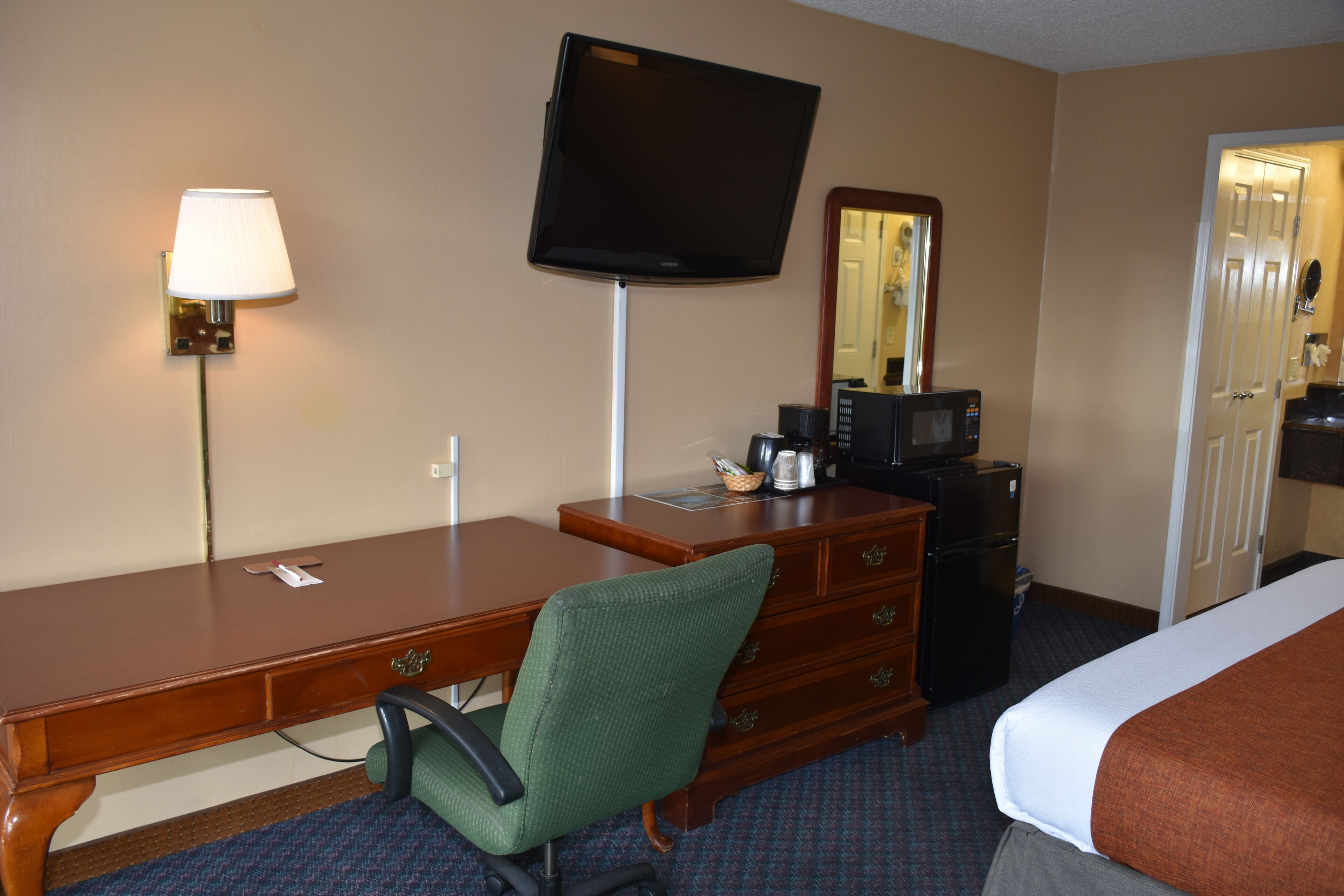 A hotel room with a wooden desk, a green chair, a television, a microwave, a refrigerator, and a bed with a brown blanket.