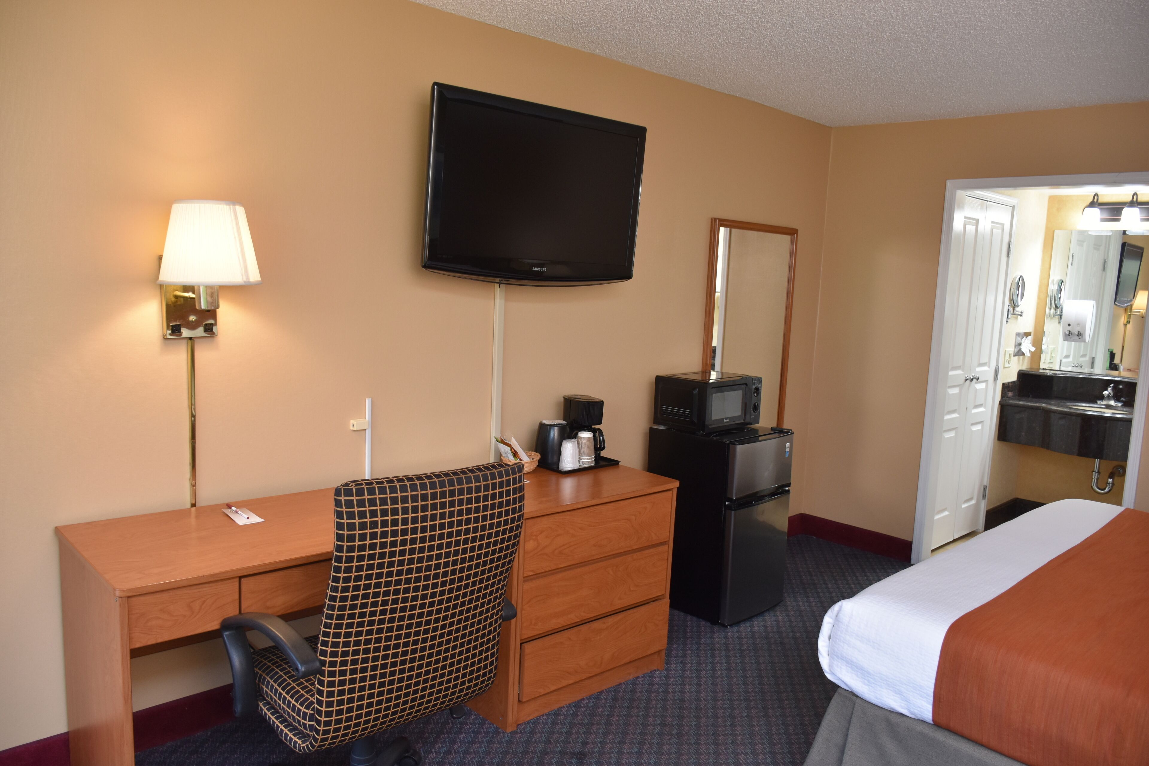 A hotel room with a bed, a desk with a chair, a television, a microwave, a refrigerator, a coffee maker, and a mirror.