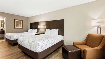 Quarto de hotel com duas camas, uma poltrona marrom, uma mesinha de apoio e uma mesa de cabeceira com abajur.